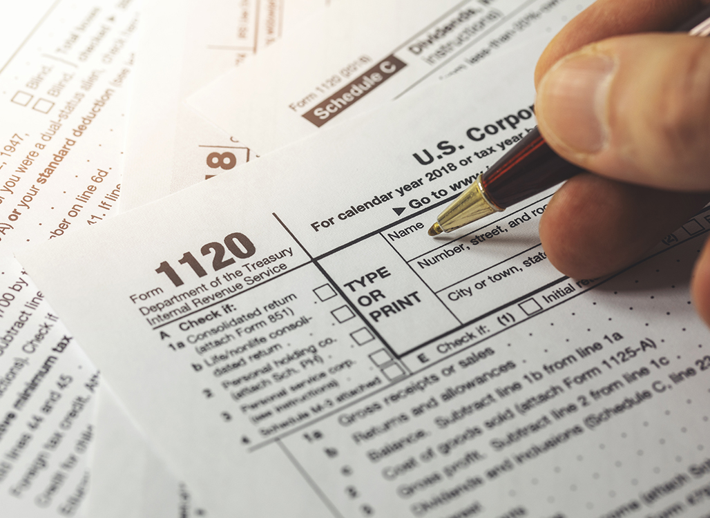 a close up image of a person's hand using a pen to fill out a corporate tax form